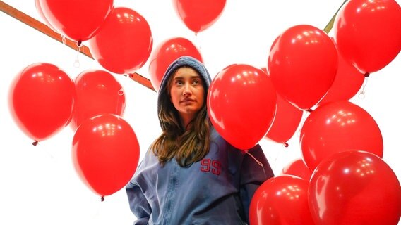 Eine junge Frau steht mit einem Kapuzenpulli vor einem weißen Hintergrund. Um sie herum fliegen rote, runde Luftballons, zu denen sie hinaufschaut. © Olaf Struck Foto: Olaf Struck