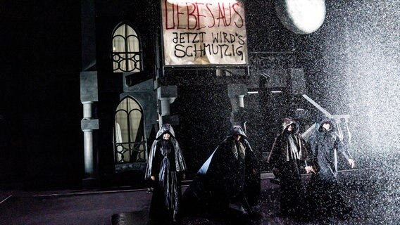 Vier Personen in dunklen Umhängen im Regen auf der Bühne im Stück "Noch wach?" im Thalia Theater © Krafft Angerer / Thalia Theater Foto: Krafft Angerer