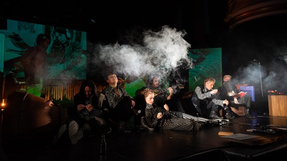 Schauspielerinnen und Schauspieler sitzen in einer Rauchwolke auf einer Bühne © Schleswig-Holsteinisches Landestheater / Henrik Matzen Foto: Henrik Matzen