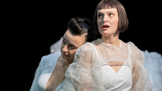 Zwei Schauspielerinnen stehen mit aufwendigen weißen Klamotten auf der Bühne. Die eine schaut traurig und die andere ist voller Tatendrang. © Oldenburgisches Staatstheater Foto: Stephan Walzl