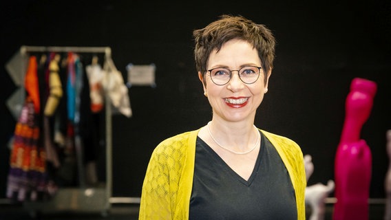 A woman with short hair, glasses and a yellow cardigan smiles openly at the camera © Staatstheater Hannover Photo: Kerstin Schomburg