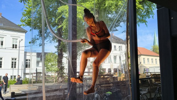 Eine Frau hängt eingeschweißt zwischen zwei durchsichtigen Folien. Sie trägt ein Top und eine kurze Hose. © Helgard Füchsel / NDR Foto: Helgard Füchsel / NDR