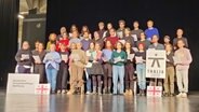 Viele Menschen stehen zu einem Chor aufgereiht auf einer Theaterbühne. © NDR.de Foto: Peter Helling