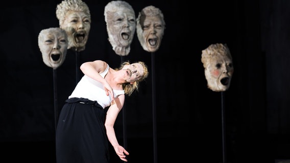Eine Frau mit Maske auf einer Bühne - im Hintergrund große Masken. © Monika Rittershaus Foto: Monika Rittershaus