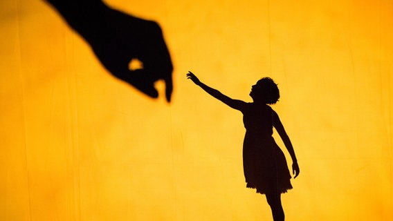 Der Schatten eines Mädchens streckt seine Hand einer großen Faust vor einer gelb beleuchteten Leinwand entgegen. © Ian Douglas 
