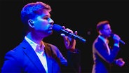 Zwei junge Männer im Anzug stehen auf der Bühne und singen ins Mikrofon © The Real Group 