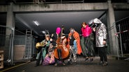Eine bunt gekleidete Gruppe steht mit Musikinstrumenten vor einem baufälligen Haus. © Andrea Vollmer 