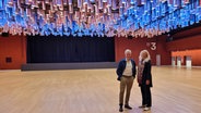 Eine Frau und ein Mann stehen in einem leeren Saal; an der Decke hängen Kannenlampen im 70er-Jahre-Design. © NDR Foto: Peter Helling