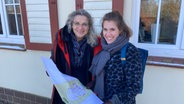 Zwei Frauen stehen mit einem Dokument vor einem Haus. © NDR Foto: Andrea Schwyzer