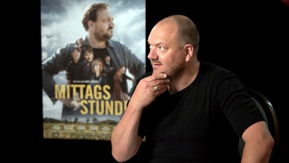 Charly Hübner vor einem Plakat für den Film "Mittagsstunde" © NDR Foto: Lornz Lorenzen