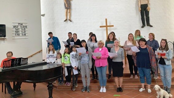 Ein Chor aus Frauen, Männern und Kindern singt, begleitet vom Chorleiter am Klavier, im Gemeindesaal der evangelischen Bonhoeffer-Gemeinde in Wolfsburg. © NDR Foto: Janek Wiechers