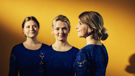 Drei junge. blonde Frauen mit hochgesteckten Haaren stehen sich teils gegenüber. Sie lächeln und tragen dunkelblaue Abendkleider. Der Hintergrund strahlt gelbgold. © Carolin Bittencourt 