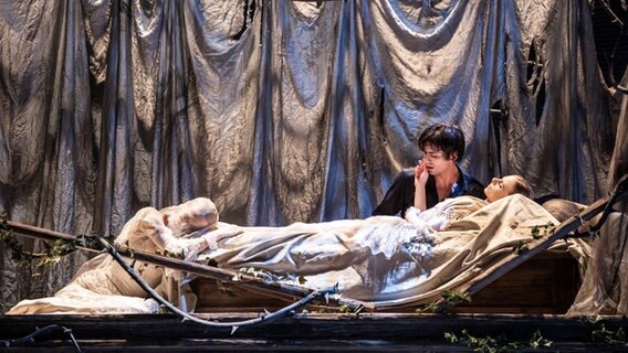 Der Prinz kniet am Bett von Dornröschen, die mit weißen Decken und Gewändern zugedeckt ist. Er sieht verzweifelt aus. © Kiran West Foto: Kiran West