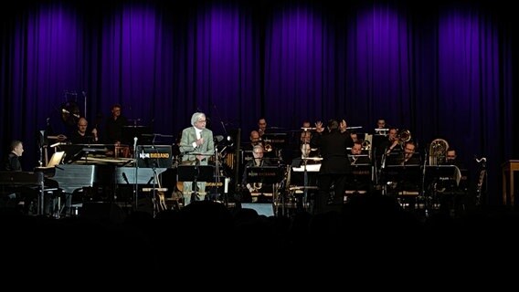 Ein Sänger mit Anzug und grauem, schulterlangem Haar steht auf einer Bühne. Hinter ihm ist ein Orchester. © NDR Foto: Pearl Väth