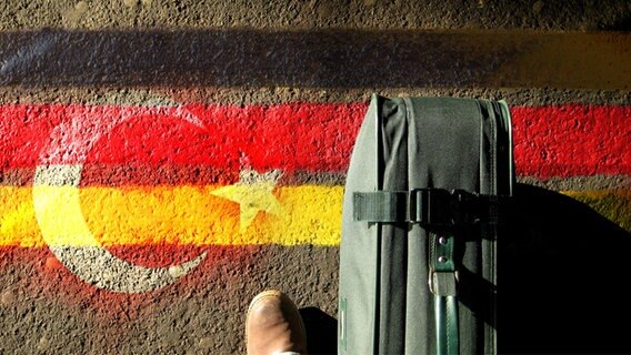 Koffer steht auf Asphalt, der mit einem Graffiti bemalt ist, das deutsche und türkische Flaggensymbole zeigt. © photocase Foto: photographe