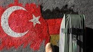 Koffer steht auf Asphalt, der mit einem Graffiti bemalt ist, das deutsche und türkische Flaggensymbole zeigt. © photocase, fotolia Foto: photographe, cil86