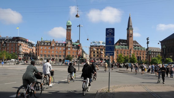 Eine breite Kreuzung mitten in einer Großstadt. Fünf Fahrradfahrer fahren Richtung der leeren Straßen. © IMAGO / Dean Pictures 