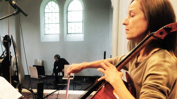 Eine Frau mittleren Alters mit langen roten Haaren spielt Cello. Im Hintergrund sieht man zwei Kirchenfenster und einen Techniker. Ein Mikrofon ist auf sie ausgerichtet. © NDR Foto: Horst Konietzny