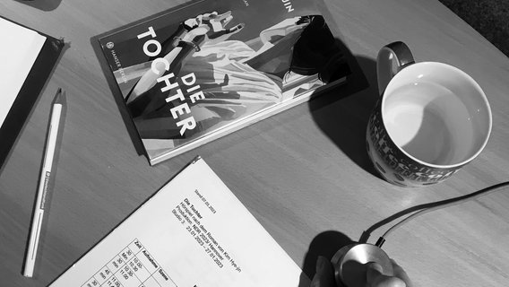 Ein Schreibtisch mit einer Tasse, einem Roman "Die Tochter", einer Mouse und einem Dokument in schwarzweiß © NDR/Alina Rohrer 