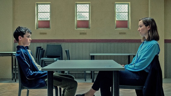 Ein Teenager-Junge mit dunklen kurzen Haaren sitzt in einem Verhör an einem Tisch mit einer Frau mit schulterlangen Haaren in einem großen Raum mit großem Tisch - Szene aus der "Netflix"-Serie "Adolescence" mit Schauspieler Owen Cooper © Courtesy of Ben Blackall/ Netflix © 2024 