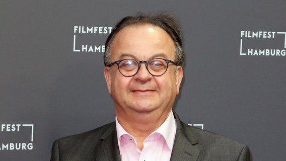 Ein lächelnder Mann im grauen Anzug steht vor einer Wand mit dem Schriftzug Filmfest Hamburg  