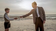 Ein Junge steht am Strand und reicht einem Mann etwas in die Hand  - Filmszene mit Matthias Schweighöfer (r.) und Jasper Billerbeck aus dem Film "Amrun" © 2025 bombero international GmbH & Co. KG / Rialto Film GmbH / Warner Bros. Entertainment GmbH / Mathias Bothor Foto: Mathias Bothor