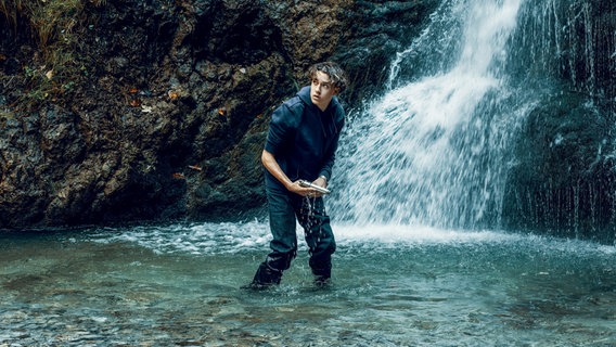 Ein Junge steht in einem Wasserbecken unterhalb eines Wasserfalls und hält eine Pistole in der Hand. © ARD Degeto Film/Bantry Bay Productions GmbH 