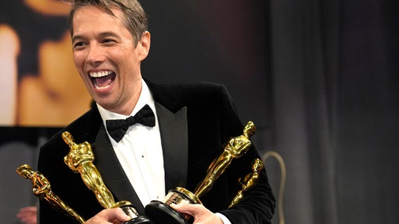 Ein Mann im schwarzen Smoking strahlt über beide Ohren und freut sich über seinen Oscar für den besten Film, dahinter steht sein elegant gekleidetes Filmteam © John Locher/Invision/AP/dpa +++ dpa-Bildfunk +++ Foto: John Locher