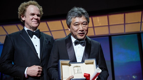 Hirokazu Koreeda (r) nimmt im Namen von Yuji Sakamoto den Preis von John C. Reilly für das beste Drehbuch für den Film "Monster" entgegen. © Daniel Cole/AP/dpa +++ dpa-Bildfunk Foto: Daniel Cole
