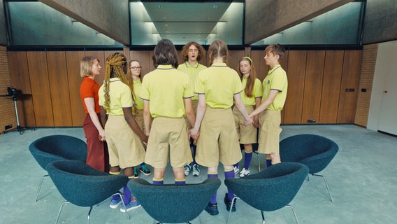 Eine Gruppe von Jugendlichen in grüngelben Uniformen steht in einem Stuhlkreis, darin eine Frau mit roter Kleidung (Szene aus "Club Zero" von Jessica Hausner) © Neue Visionen / dpa Bildfunk 