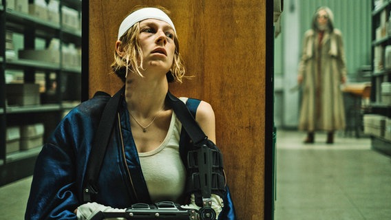 Eine Frau mit verletztem Kopf (mit einer weißen Binde), einer ARmschiene und lädiertem Gesicht hockt hinter einem Buchregal - Szene aus dem Horrorfilm "Cuckoo" © Weltkino Verleih 