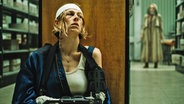 Eine Frau mit verletztem Kopf (mit einer weißen Binde), einer ARmschiene und lädiertem Gesicht hockt hinter einem Buchregal - Szene aus dem Horrorfilm "Cuckoo" © Weltkino Verleih 