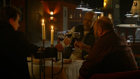 Drei Männer mittleren Alters prosten sich in einer Bar mit vollen Rotweingläsern zu. (Ausschnitt aus "Eine Nacht in Helsinki" von Mika Kaurismäcki.)  