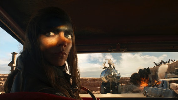 Eine Frau sitzt im dunklen Auto, nur ihre Augen sind hell erleuchtet - im Hintergrund ein Tankwagen - Szene aus "Furiosa" © 2023 Warner Bros. Entertainment Inc. All Rights Reserved. Foto: Jasin Boland