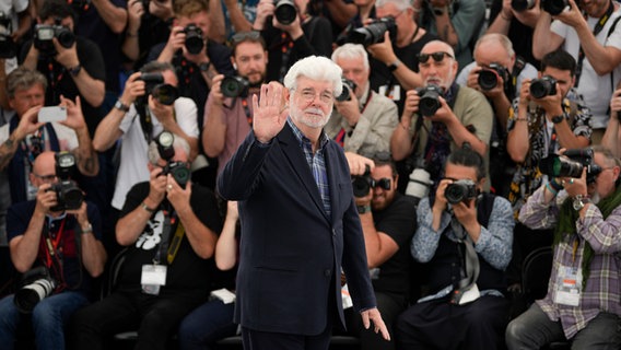 Ein Mann mit weißem Bart und Brille winkt den Fotografen - US-Filmemacher George Lucas bei einem Pressetermin beim Filmfest Cannes ©  Andreea Alexandru/Invision/AP/dpa +++ dpa-Bildfunk ++ Foto:  Andreea Alexandru