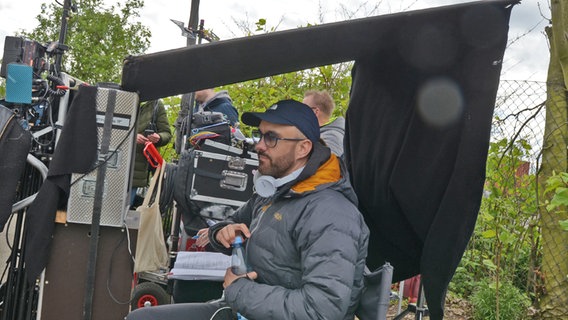 Ein Mann mit Brille und dunkler Käppi sitzt unter einem kleinen Zeltdach - der Regisseur Florian Gottschick bei Dreharbeiten am Hafen zum Spielfilm "Triage" der Serie "Großstadtrevier" © NDR Foto: Patricia Batlle