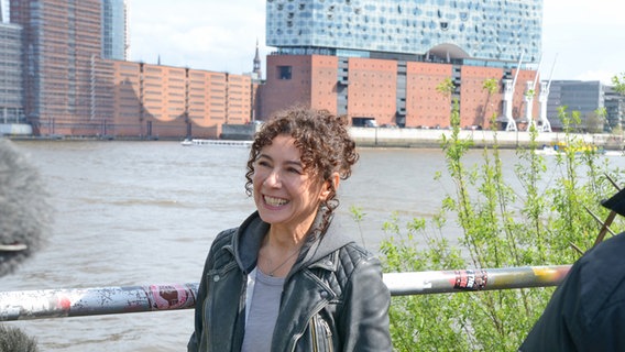 Eine lächelnde Schauspielerin mit dunklen Haaren und Locken (Maria Ketikidou) lächelt am ´Hafen von Hamburg, im Hintertgrund die Elbphilharmonie © NDR Foto: Patricia Batlle