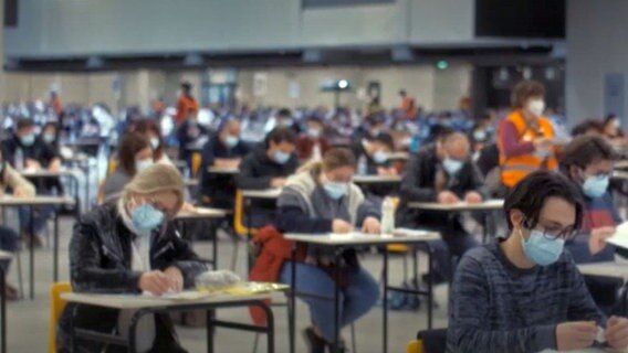 In einer großen Halle sind in regelmäßigen Abständen Tische verteilt, an denen Menschen sitzen, wie in einer Prüfungssituation. © Screenshot Il Posto 