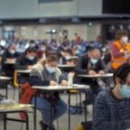 In einer großen Halle sind in regelmäßigen Abständen Tische verteilt, an denen Menschen sitzen, wie in einer Prüfungssituation. © Screenshot Il Posto 