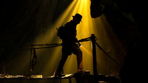 Harrison Ford als Indiana Jones leuchtet den Weg in einer Grotte mit gelbem Licht mit einer Taschenlampe - Szene aus "Indiana Jones und  der Ruf des Schicksals" © 2022 Lucasfilm Ltd. & TM 