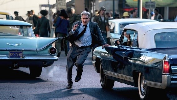 Ein Mann mit Krawatte rennt zwischen Autos auf einer Straße - Szene mit Harrison Ford aus "Indiana Jones und das Rad des Schicksals" © 2022 Lucasfilm Ltd Foto: Jonathan Olley
