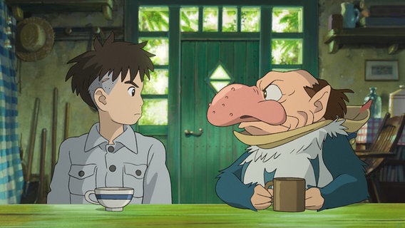Ein Junge und ein Wesen mit einer sehr großen menschlichen Nase sitzen nebeneinander an einem grünen Tisch und blicken sich ernst an - Szene aus "Der Junge und der Reiher" vom Studio Ghibli (Hayao Miyazaki) © © STUDIO GHIBLI 2023 