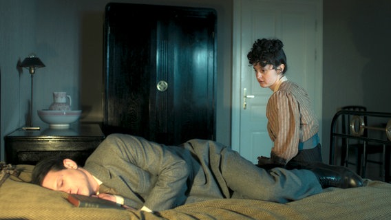Kafkas Schwester Ottla (Maresi Riegner) sitzt am Bett neben ihrem liegenden Bruder in der ARD-Serie "Kafka". © NDR/Superfilm 