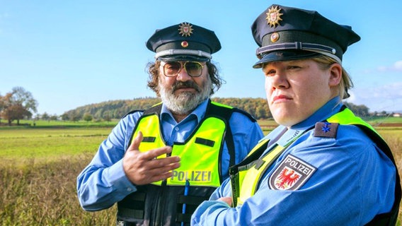 Eine Polizistin und ein Polizist mit gelben Warnwesten schauen grimmig in die Kamera - Szene aus dem Roadmovie "Kannawoniwasein!" © Weltkino 