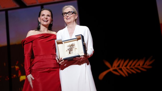 Juliette Binoche (links) im roten Kleid und Meryl Streep im weißen Kleid lachen auf der Bühne des Filmfestivals Cannes, wo die Schauspielerin Streep die Goldene Ehrenpalme erhalten hat © Vianney Le Caer/Invision/AP/dpa +++ dpa-Bildfunk +++ Foto: Vianney Le Caer