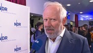 Ein Mann mit blauen Augen, weißen Haaren und dunklem Anzug spricht in einem Interview (Axel Milberg) © NDR Screenshot 