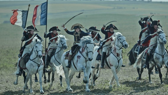 Der Heerführer Napoleon reitet mit weiteren Soldaten und der französischen Fahne auf einem Schlachtfeld - Szene mit Joaquin Phoenix aus "Napoleon" von Ridley Scott © AppleTV+ 