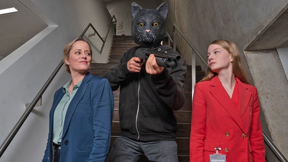 Zwei Frauen auf einer Treppe, dahinter eine Person mit Katzenkopf und Gewehr - eine Szene der Serie "Die nettesten Menschen der Welt" © NDR/Studio Zentral/Michael Ihle Foto: Michael Ihle