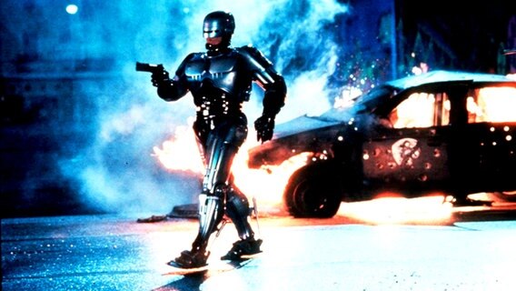 Filmstill aus "RoboCop" mit Peter Weller als Robocop mit einem Roboter als Polizisten, der aus einem brennenden Auto steigt © Picture-Alliance / Photoshot 