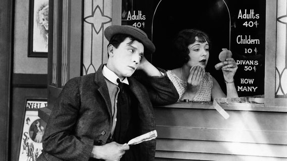 Buster Keaton steht an einer Kinokasse, die Kinokartenverkäuferin  pudert sich die Nase - Szene von 1924 aus dem Stummfilm "Sherlock, Jr." von Buster Keaton © picture alliance / Everett Collection | Courtesy Everett Collection 